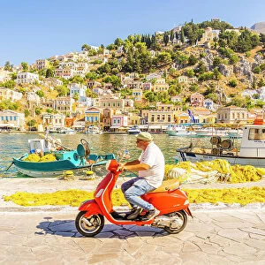 A local man riding a red Vespa in the colourful harbour in Symi, Dodecanese Islands, Greece