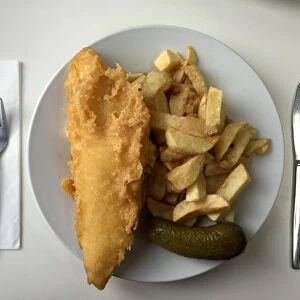 A plate of traditional fish and chips with a gherkin on a table in a seafront fish