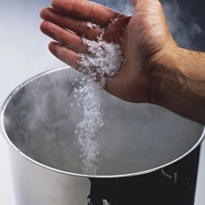 Hand pouring coarse salt grains into steaming saucepan, elevated view