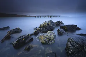 Tranquility Prints: Stormy weather with heavy rain clouds at bay with rocks and lichen at foreground and old pier
