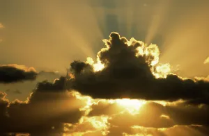 Low Angle View Prints: Sun rays beam from behind cumulus clouds