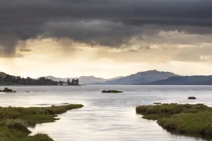 View of meandering river flowing through grassland with hills at background and rain clouds